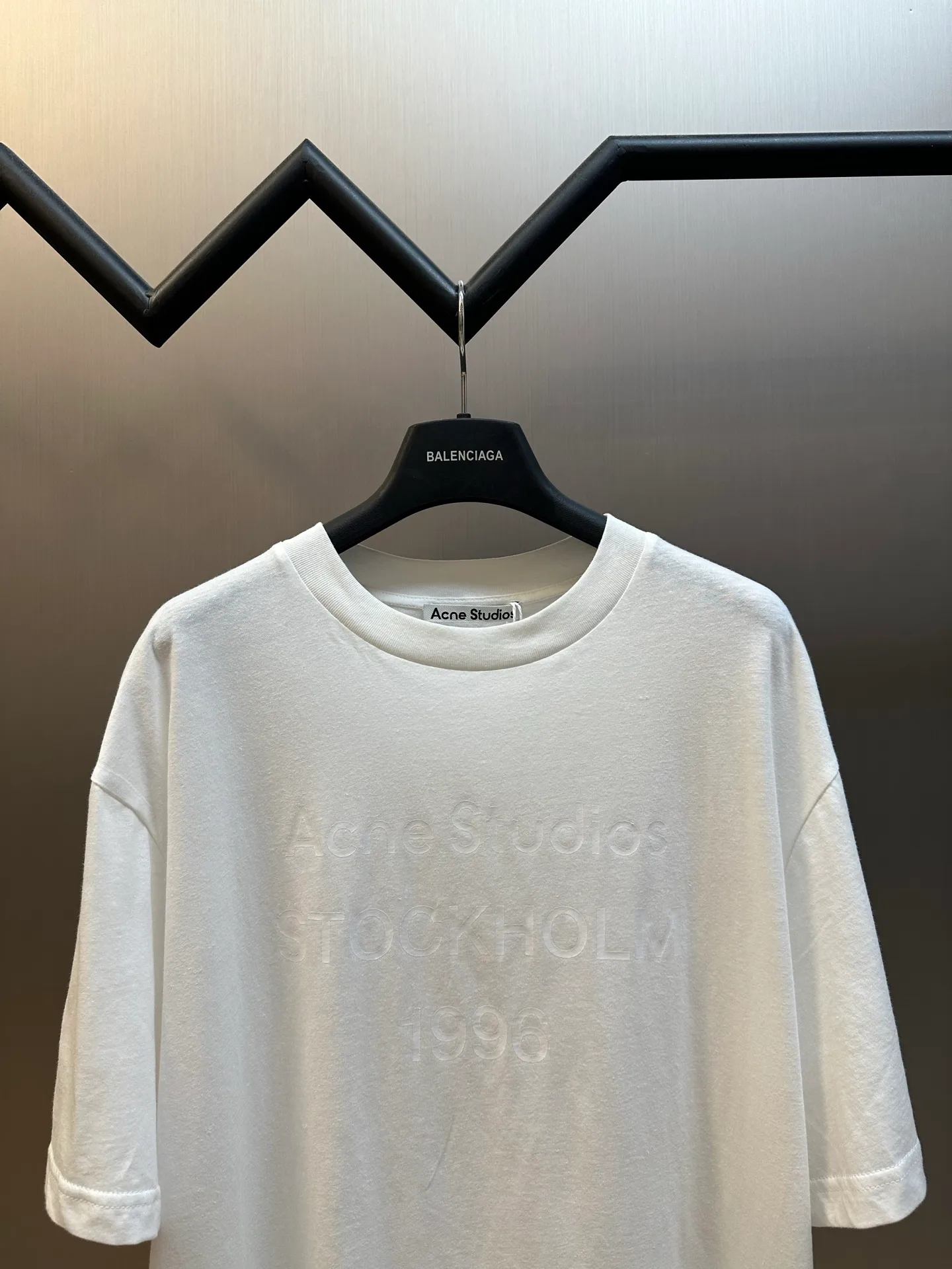 Ac White Letter Round Neck Short Sleeve The fabric is made of 32-count cotton and linen blended plain knitting, with a ratio of 85% cotton and 15% linen fiber. The weight is only 145g/m²; the ribbed neckline is made of Same Style cotton and linen blended yarn, 145g/m², to ensure a unified texture of the entire piece. The collision of the natural rough base and the industrial spraying process creates a natural irregular texture and an extremely light and breathable wearing experience. Cotton provides a skin-friendly and soft foundation, while linen introduces natural roughness and knotted particles, giving the fabric surface an irregular texture. The addition of hemp fiber makes the fabric have excellent moisture absorption and perspiration properties, while also bringing a natural micro-wrinkle texture and dry touch. After the fabric is piece-dyed, the dye evenly penetrates into the fiber, showing full color fastness and soft matte color, providing a uniform base for subsequent inkjet printing. The front is decorated with logo printing. The printing uses a double-layer printing process of environmentally friendly elastic glue and varnish, which takes into account the concept of environmental protection, wearing comfort and visual expression. The bottom layer uses environmentally friendly elastic glue, with water-based environmentally friendly resin as the base material. The color paste is scraped onto the surface of the fabric through a high-mesh screen. After drying and solidifying at high temperature, a soft and highly elastic continuous adhesive film is formed. The printed film can stretch and retract with the fabric. Even if it is covered with a large area of ​​pattern, it is still breathable and does not feel tight when wearing it. The surface layer is accurately overprinted with transparent varnish, creating a high-gloss effect in specific areas. This layer of varnish not only significantly improves the color contrast of the pattern, but also creates a local wet and bright visual and tactile contrast, making ordinary printing instantly gain three-dimensional layers and glossy texture. At the same time, the smooth surface layer also enhances Wear-Resistant and wash-resistant properties. This T-shirt is defined as a neutral style, casual fit, and hip length; its style logic pursues a genderless sense of wrapping and extended proportions. The key point is that the dress is hip-length, not only to cover the buttocks, but also to create a lazy, flowing silhouette by utilizing the lengthened body and the drape of the fabric. In terms of process execution, the side seams need to maintain a straight hem to prevent the long body from looking like a straight bucket. Taken as a whole, it's a hybrid that combines the functional details of workwear with the relaxed feel of high fashion. size：xs s m l